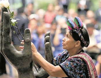 Una mujer de perfil con traje tradicional indígena y tocado colorido coloca una rosa blanca en una escultura de grandes manos oscuras, con gente difuminada detrás