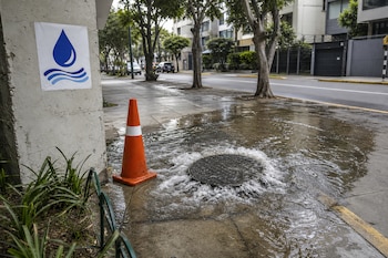 Agua saliendo de una tapa de alcantarillado en una vereda junto a un cono de señalización naranja y un cartel con el símbolo de una gota.