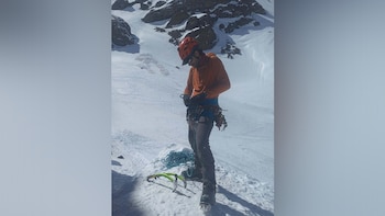 Un hombre con casco naranja y arnés de escalada manipula una cuerda sobre la nieve en una ladera montañosa