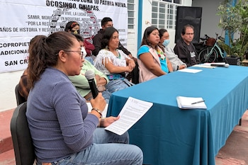 Una mujer habla por micrófono sosteniendo un documento frente a una mesa con mantel azul; detrás, varias personas sentadas y un banner con texto