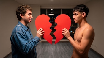 Dos hombres jóvenes sostienen frente a frente sendas mitades de un corazón rojo recortado en un interior con ventanal al fondo.