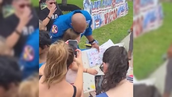 Un astronauta de uniforme azul abraza a un niño en un evento de bienvenida. Varias personas observan y graban. Al fondo, carteles con banderas de EE.UU