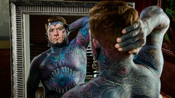 Un hombre con el torso, el cuello y el rostro cubiertos por tatuajes de colores mira hacia un espejo de marco plateado