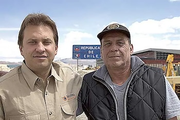 Dos hombres, uno con camisa y otro con gorra y chaleco, posan frente a un cartel azul que dice "República de Chile" y maquinaria amarilla