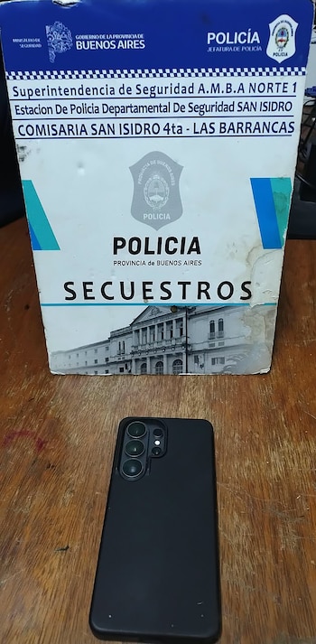 Un teléfono celular negro sobre una mesa de madera frente a un cartel blanco de la Policía de la Provincia de Buenos Aires