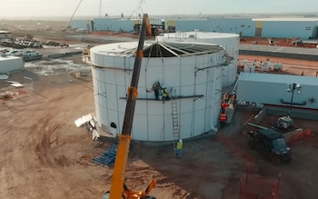 Vista aérea de un sitio de construcción con un gran tanque cilíndrico blanco en primer plano, trabajadores en andamios, una grúa y terrenos marrones