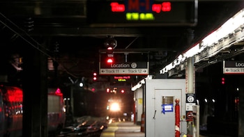 Tren con luz frontal brillante se acerca a un andén de metro oscuro; luces rojas de seguridad, señales de ubicación y vías son visibles en la estación
