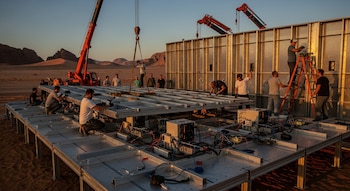Vista de científicos ensamblando una estructura metálica gigante con paneles de aluminio y equipos tecnológicos bajo grúas en un paisaje desértico al atardecer.