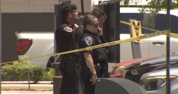 Tres oficiales de policía uniformados observan detrás de una cinta amarilla de "Crime Scene Do Not Cross" con vehículos y árboles de fondo