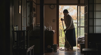 Una persona barre hojas secas del suelo de madera en una entrada interior de una casa. La luz natural entra por una ventana, revelando un ambiente hogareño.
