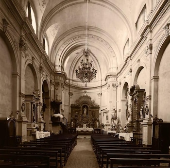 Vista interior de una iglesia con techo abovedado, altar principal, bancas de madera, candelabro central y altares laterales con estatuas