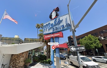 Letrero azul y blanco de Mel's Drive-In con la figura de un pingüino, situado junto a una calle con tráfico y una bandera estadounidense