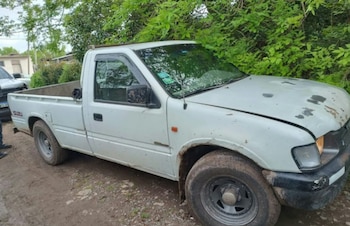 Una camioneta blanca con restos de barro estacionada junto a árboles en un camino de tierra