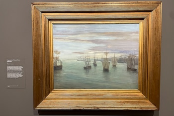 Una pintura en un marco dorado ornamentado muestra un puerto con múltiples barcos de vela bajo un cielo nublado, expuesta en una pared gris