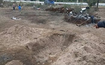 Hoyo en un terreno de tierra con montículos de arena, escombros al fondo, dos niños a lo lejos y una mano que señala el suelo
