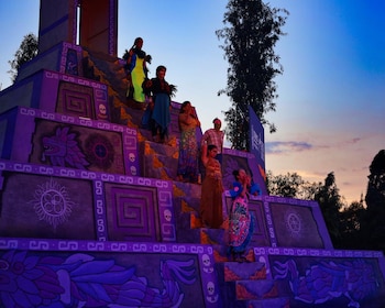 Varias personas con trajes coloridos están de pie sobre una pirámide morada con motivos grabados durante el atardecer