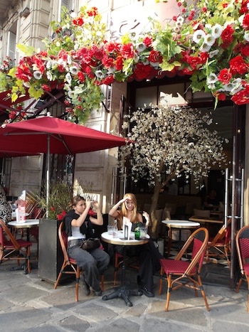 Dos personas están sentadas a la mesa de la terraza de un café al aire libre decorado con flores rojas y blancas