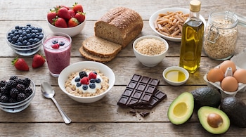 Una mesa de madera con cuencos de arándanos, frutillas, moras, arroz, pasta, avena, pan integral, batido, huevos, aguacate, chocolate negro y aceite de oliva.