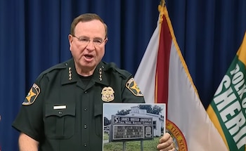 Un hombre vestido con uniforme de sheriff sostiene la fotografía de una iglesia con un cartel; detrás, banderas del estado de Florida y de la oficina del Sheriff de Polk