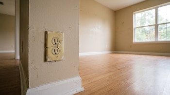 Una toma de corriente beige agrietada en primer plano sobre una pared, con un suelo de madera y una ventana al fondo.