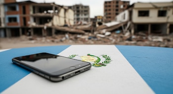 Un smartphone negro sobre una superficie que representa la bandera de Guatemala. Al fondo, edificios de hormigón con daños estructurales y escombros.