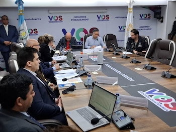 Personas en una mesa de conferencia con laptops y micrófonos, frente a banderas y pancartas del grupo político VOS Bancada Guatemala. Una bandera de Guatemala es visible