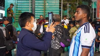Un funcionario con mascarilla fotografía con una tableta el rostro de un hombre frente a otras personas y agentes policiales