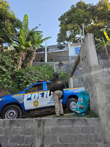 Un policía de la DEIC de espaldas junto a una patrulla azul de la PNC. Otros agentes en cascos negros se ven en balcones de una casa blanca en una colina