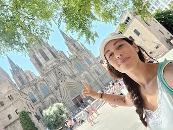 Una mujer con gorra blanca y blusa de tirantes señala hacia una catedral gótica de piedra en una plaza iluminada por el sol