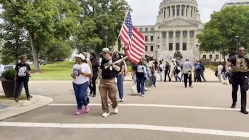 Grupo de personas, una de ellas con una bandera de Estados Unidos, marchando por una calle pavimentada. Al fondo se ve un edificio con cúpula y árboles