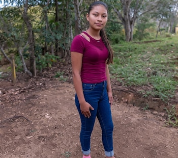 Una mujer joven de pie sobre un camino de tierra, vestida con camiseta de color magenta y pantalones vaqueros azules, con árboles al fondo