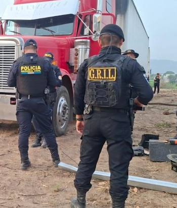 Dos agentes policiales de espaldas, uno con chaleco de Policía Nacional Civil y otro de GRIL, frente a un camión rojo con remolque blanco en un terreno de tierra