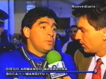 Primer plano de Diego Maradona con camiseta azul de Boca Juniors, hablando ante un micrófono verde con logo de NueveDiario. Un periodista lo entrevista