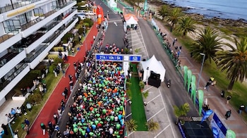 Vista aérea de una multitud de corredores en la línea de salida de una carrera, con un arco que indica 5k, 10k, 21k y 42k, carpas, un edificio de apartamentos, palmeras y el océano