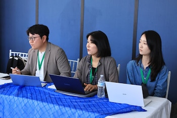 Tres personas con credenciales verdes sentadas en una mesa con tres laptops, un teléfono móvil, una botella de agua y una pluma. Mesa cubierta con tela azul