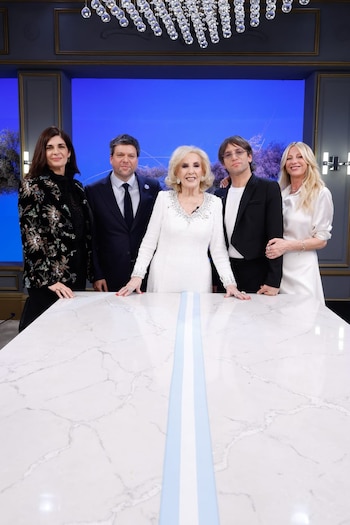 Cinco personas de pie detrás de una mesa blanca con una franja celeste y blanca. Mujer con vestido blanco en el centro. Fondo azul y araña de cristal