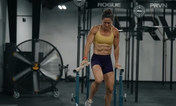 Tia-Clair Toomey, con top deportivo amarillo y shorts morados, se apoya en barras paralelas en un gimnasio con un ventilador grande de fondo
