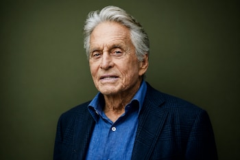 Michael Douglas luce una camisa azul desabotonada y una chaqueta oscura sobre un fondo verde.