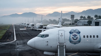 Una fila de aviones blancos en la pista de un aeropuerto, con un sello oficial en el fuselaje delantero, torre de control y montañas detrás.