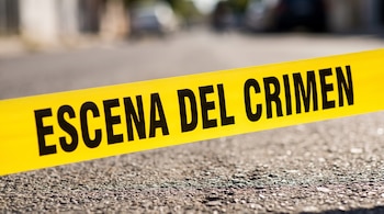 Una cinta amarilla con el texto "Escena del crimen" en letras negras mayúsculas atraviesa la parte inferior de la imagen sobre asfalto gris texturizado.