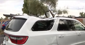 Un vehículo blanco con el techo hundido y la ventanilla trasera lateral rota, estacionado frente a árboles y postes eléctricos