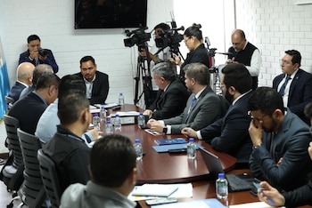 Hombres sentados alrededor de una mesa de conferencia en una sala blanca con ladrillos, con cámaras de video y un fotógrafo en la parte trasera
