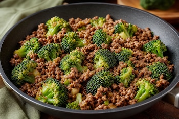 Carne picada cocida y trozos de brócoli verde distribuidos en una sartén negra sobre una superficie de madera.
