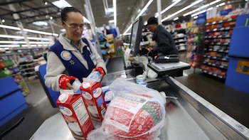 Cajera de Walmart con gafas y chaleco azul escanea cartones de crema y un pavo en una caja registradora. Otra persona trabaja al fondo