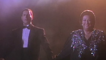 Freddie Mercury en esmoquin y Montserrat Caballé con un vestido negro bordado sonríen en un escenario iluminado