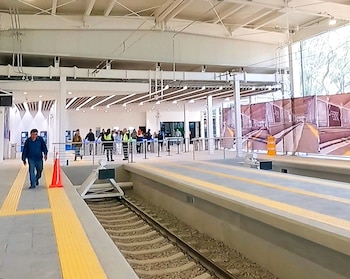 Estación de Tren Ligero Tasqueña con techos altos, andenes grises con líneas amarillas, y vías. Personas caminan, incluyendo personal con chalecos