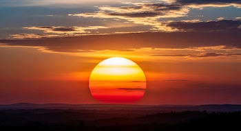 Sol grande y brillante en el centro de un cielo atardecer, con tonos naranjas y rojos sobre siluetas oscuras de colinas y bosques.