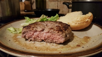 Una hamburguesa cortada por la mitad con interior rosado, un trozo de pan de hamburguesa, hojas de lechuga y grasa en un plato.