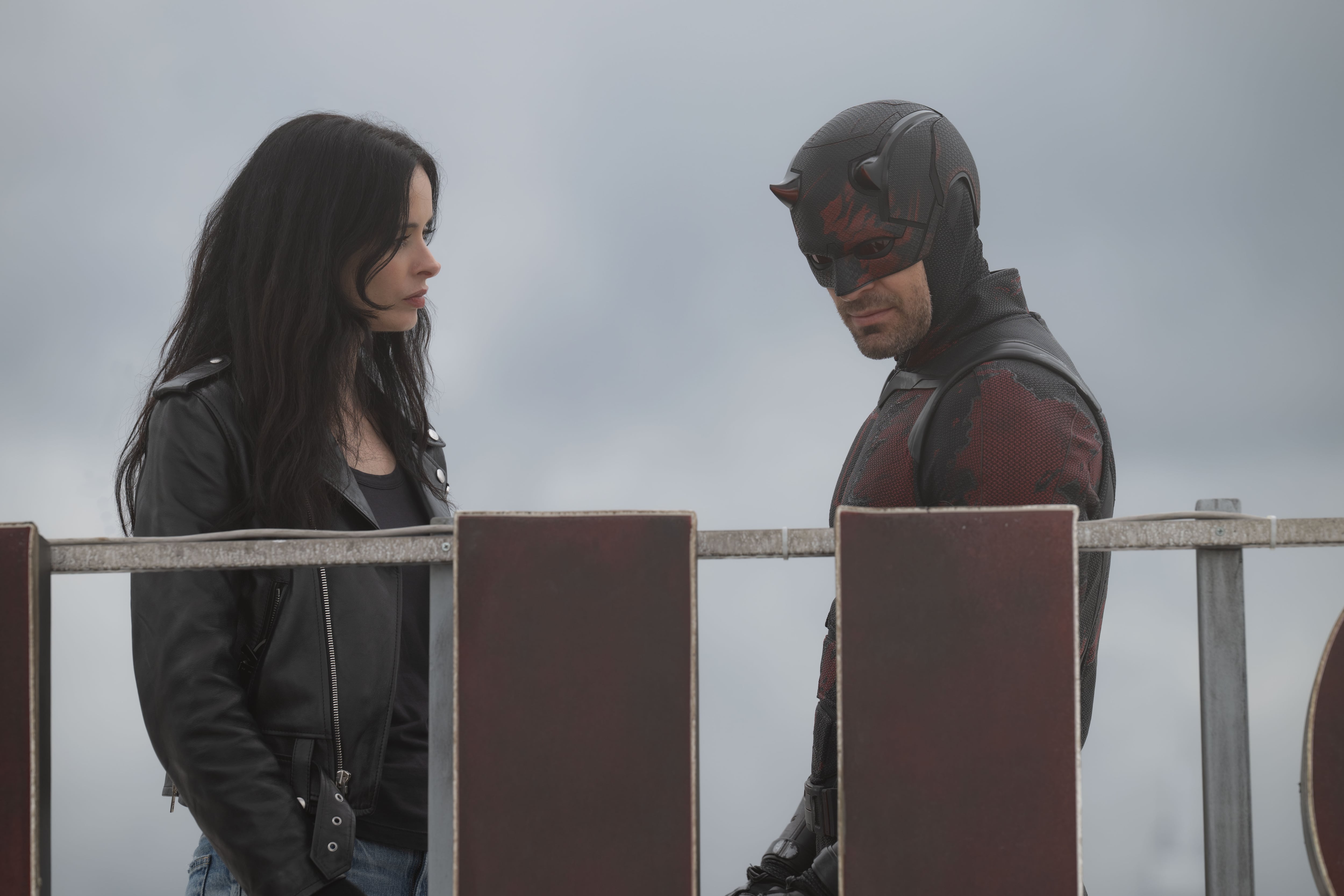 Krysten Ritter como Jessica Jones y Charlie Cox como Daredevil interactúan con semblante serio sobre un tejado bajo un cielo nublado. (disney plus)