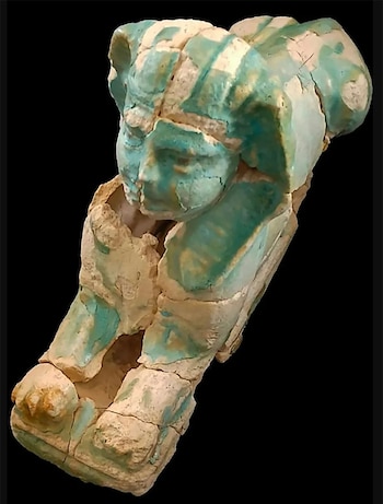 Fragmento de una escultura de un ser con cabeza humana y cuerpo animal, de colores beige y turquesa, sobre un fondo negro. La pieza está dañada